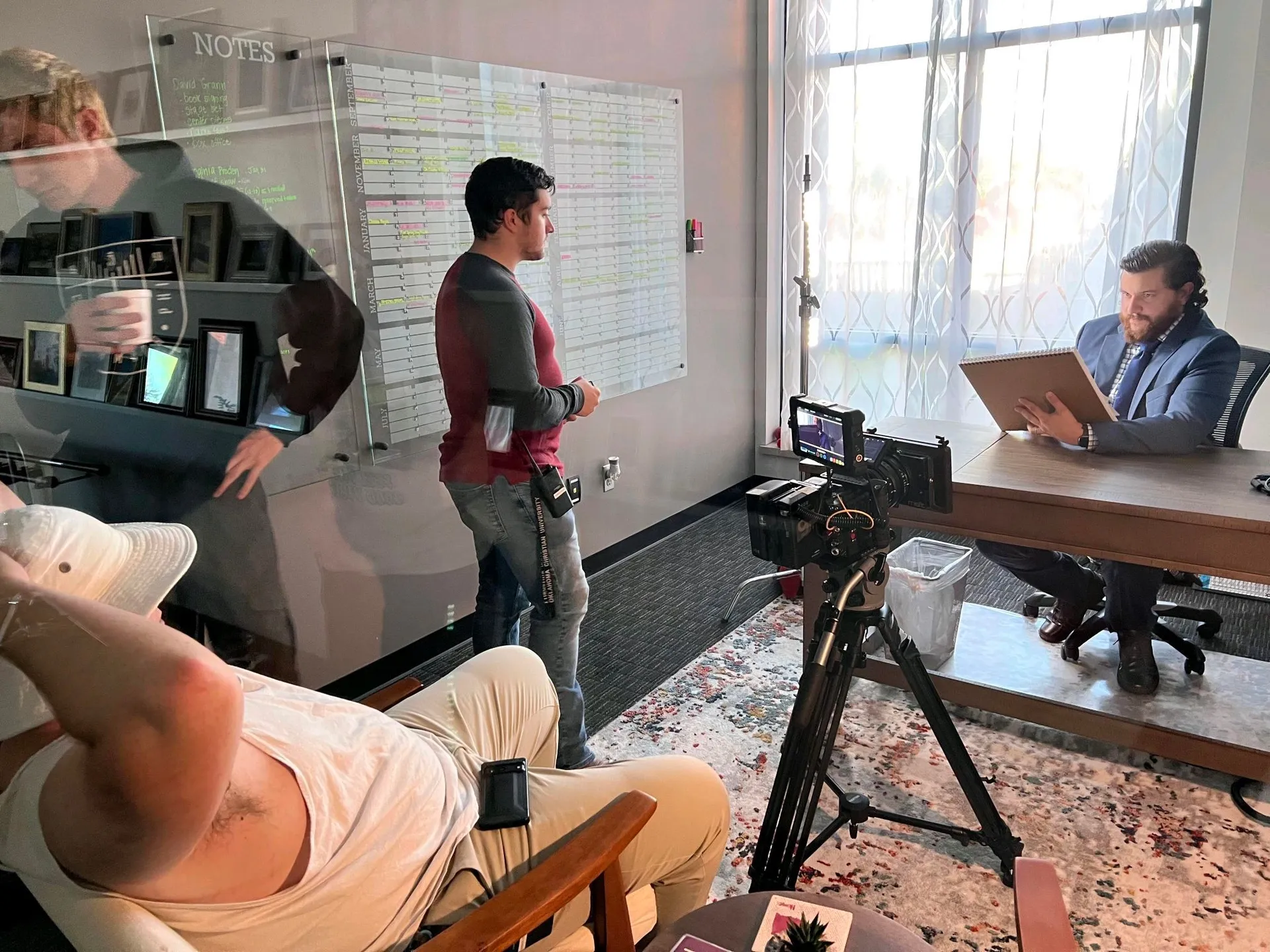 Subject in a suit reviews notes at a desk during a corporate interview shoot, with a crew member, lights, and monitor visible