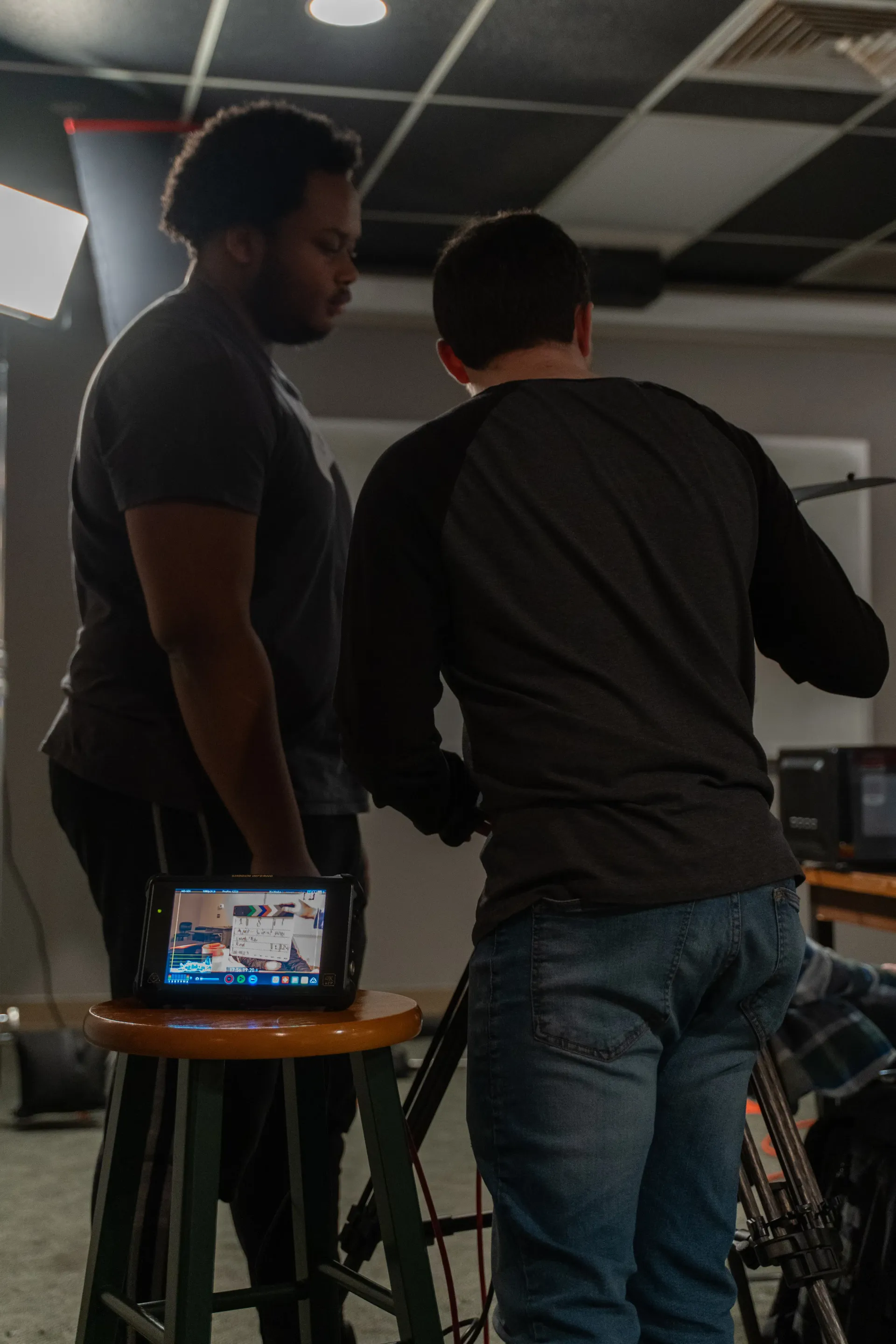 Two crew members stand behind a field monitor showing color bars and a shot on a dark set
