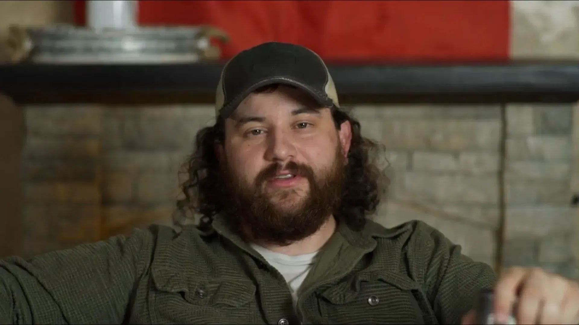 Bearded man in a cap and olive shirt speaks during an interview in front of a stone fireplace